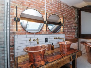 Un baño con pared de ladrillo, dos espejos redondos, tocador de madera y lavamanos de cobre.