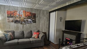 Un salón con sofá gris, cojines de colores, alfombra estampada, chimenea y televisor montado en la pared.