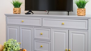 A grey cabinet with a flat-screen TV on top, two potted plants, and a white ottoman with a tray of greenery.