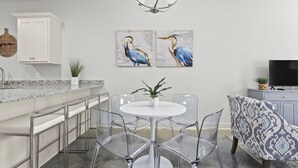 A modern kitchen with a white cabinet, a marble countertop, a round dining table with clear chairs, and two wall-mounted bird paintings.