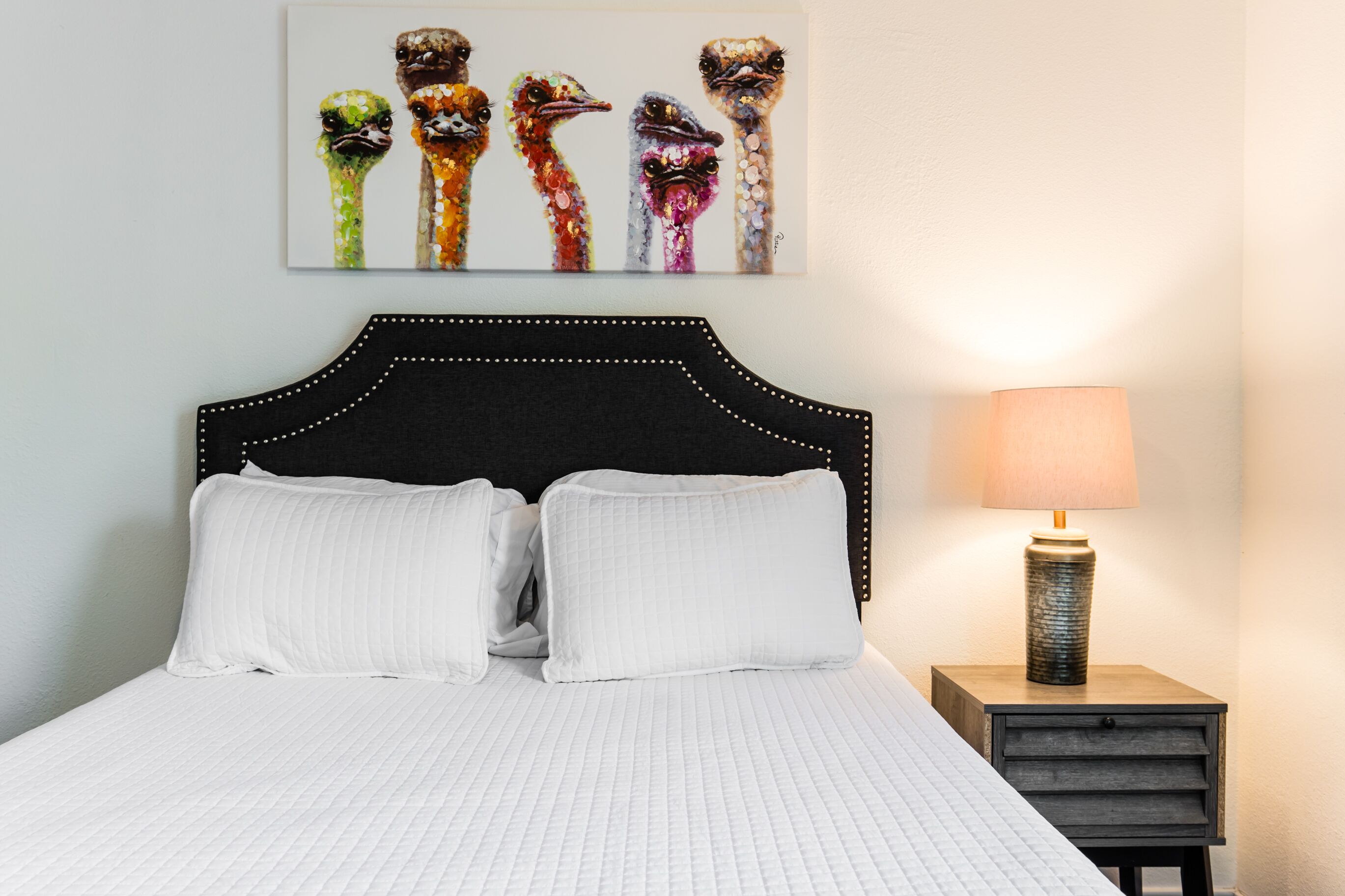 A neatly made bed with white bedding, a black headboard, and a wall-mounted painting of colorful bird heads.