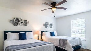 A bedroom with two beds, a ceiling fan, wall art, and a window with blinds.