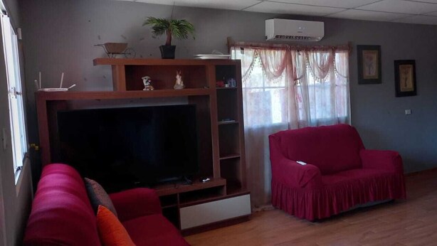 A living room with a flat-screen TV, two red armchairs, a wooden entertainment unit, and a window with curtains.