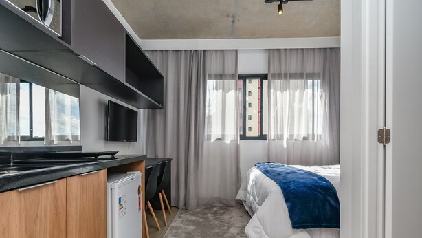 A compact hotel room with a kitchenette, a bed with a blue blanket, and a window with sheer curtains. - Vossa Bossa VN Ueno (São Paulo)