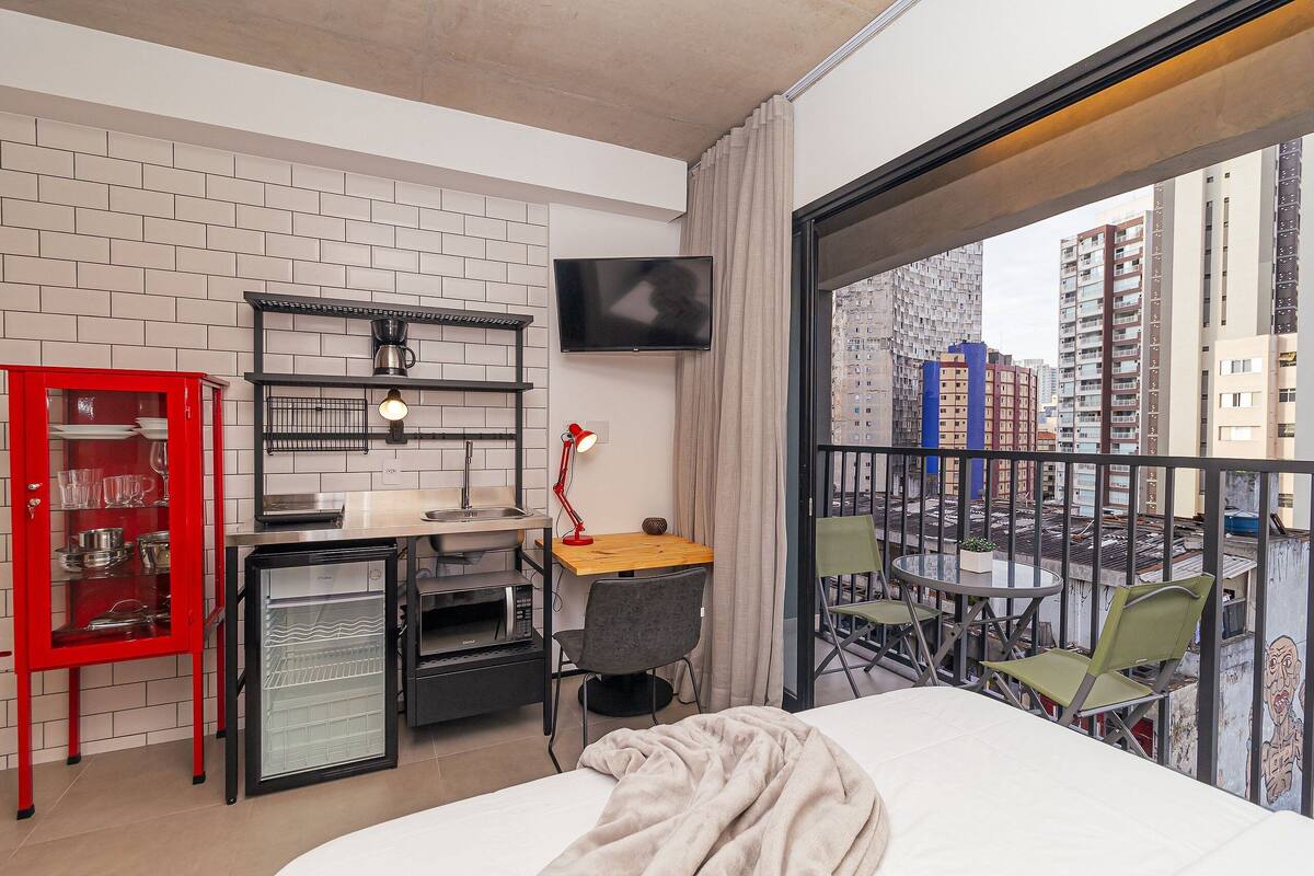 A compact hotel room with a kitchenette, a bed, a desk with a chair, a TV, and a balcony with a view of the city.
