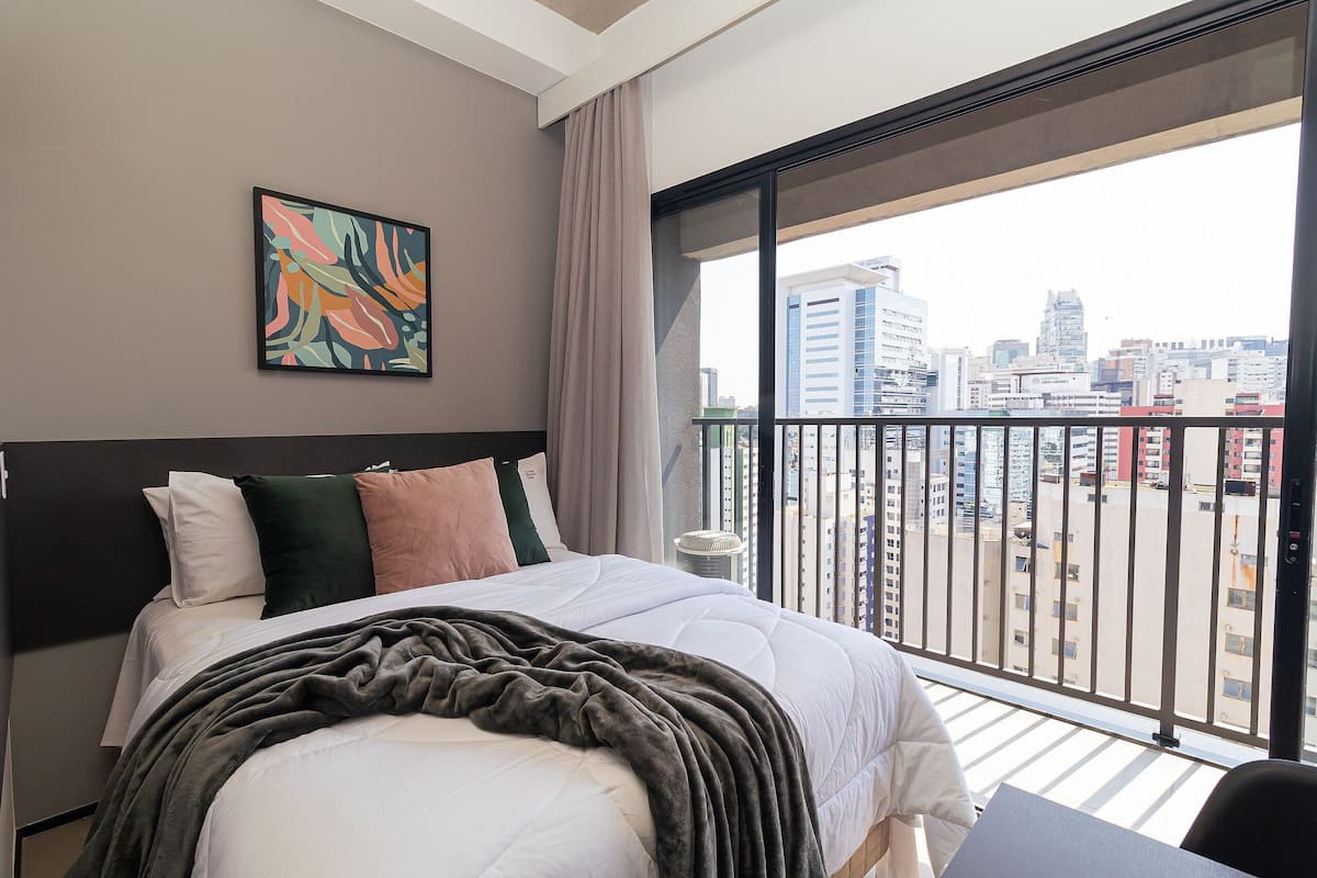 A modern hotel room with a large bed, a city view balcony, and a painting on the wall.