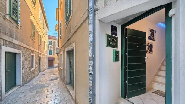 Un carrer estret amb edificis a ambdues bandes, una porta ben visible amb la inscripció “POETRY HOUSE” i una escala que mena a un pis superior.