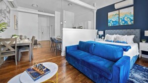 Ein modernes Hotelzimmer mit einem blauen Sofa, einem Bett mit weißer Bettwäsche, einem Essbereich mit Tisch und Stühlen sowie einer Küchenzeile.