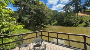 A wooden deck with metal chairs and a table overlooking a calm river, surrounded by lush greenery and trees. - Refúgio Brisa Serrana - Urubici - SC (Urubici)