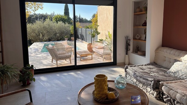 Un vaste salon doté d’une grande porte-fenêtre coulissante donnant sur une terrasse avec piscine, des meubles d’extérieur et une vue sur le jardin environnant.