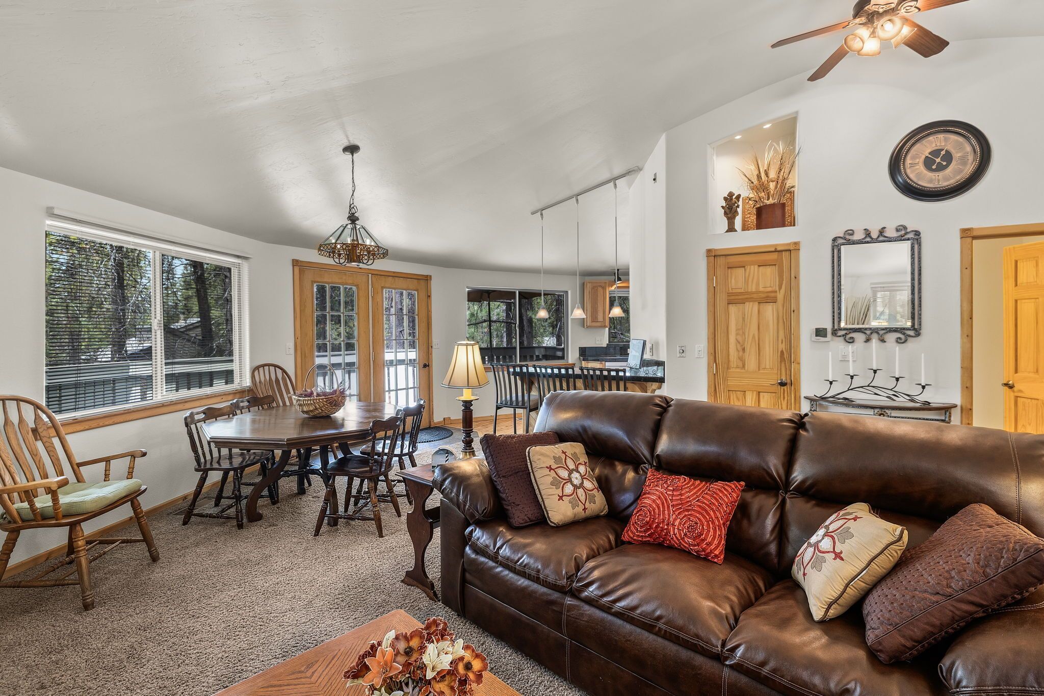 A cozy living room with a leather sofa, wooden dining table, and chairs. There is a chandelier, a clock, and a mirror on the wall.