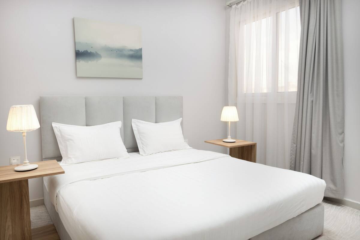 A hotel room with a large bed, two bedside tables with lamps, a headboard, a window with curtains, and a wall-mounted painting.