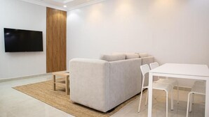 A modern living room with a sofa, a flat-screen TV, a dining table with chairs, and a wooden accent wall. - Badrani B - Qaswarah Residence  (Madinah)