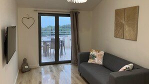 A modern living room with a grey sofa, a flat-screen TV, a ceiling light fixture, a heart-shaped decoration, and a view through a sliding glass door to an outdoor area with a table and chairs. - Appartement Idéal Famille, Belle VUE Mer, Plage du Capelan Accessible à Pieds! (Bandol)