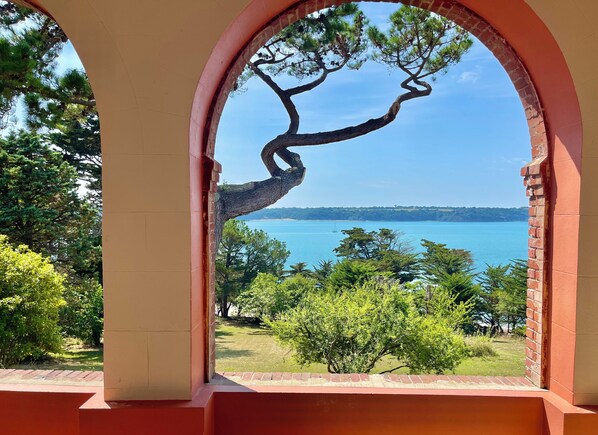 A view through an arched window overlooking a coastal landscape with a prominent tree and a clear blue sea. - Ideal for 2 families, large charming house, sea view, direct access to the beach (Saint-Jacut-de-la-Mer)