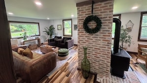 A cozy living room with a brick accent wall, a fireplace, and a large window overlooking greenery. - Charming cozy RIVERFRONT cottage on the Piney River. A little slice of heaven! (Roseland)
