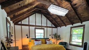 A bedroom with a wooden ceiling, a bed with yellow bedding, a large window, and a patterned rug. - Charming cozy RIVERFRONT cottage on the Piney River. A little slice of heaven! (Roseland)