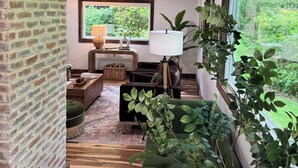 A cozy living room with a brick wall, a large window, a green sofa, a lamp, and a plant. - Charming cozy RIVERFRONT cottage on the Piney River. A little slice of heaven! (Roseland)