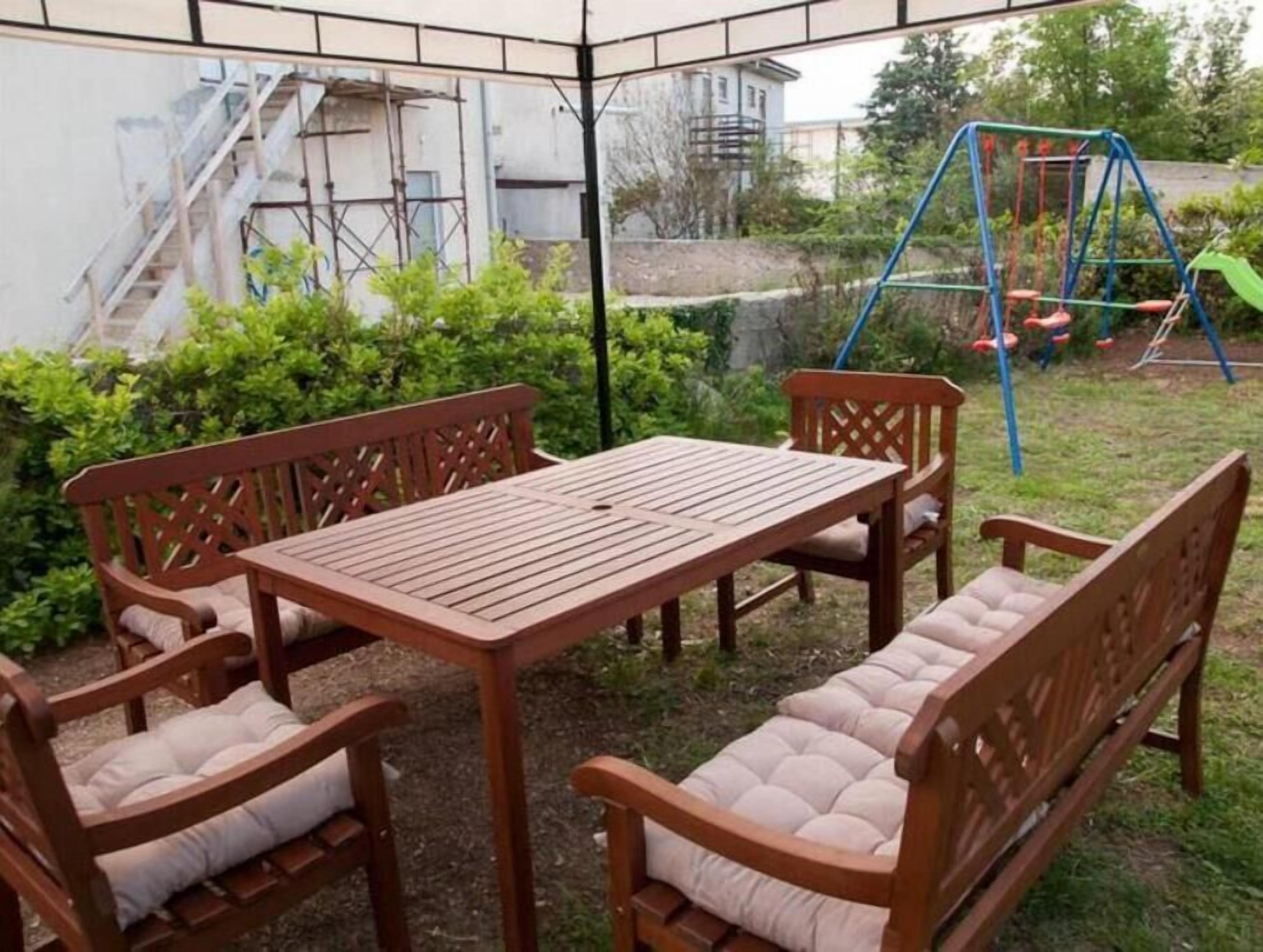 An outdoor patio with a wooden table and chairs, surrounded by greenery and a swing set.