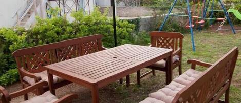 An outdoor patio with a wooden table and chairs, surrounded by greenery and a swing set.