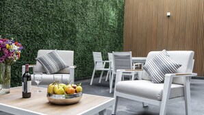 Un espacio al aire libre moderno con mobiliario blanco, una mesa de madera con un frutero y un muro de seto verde.
