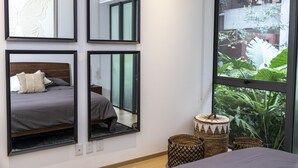 Un dormitorio con un ventanal grande, una cama con colcha gris, varios espejos y vistas a zonas verdes.