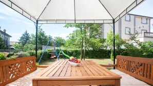 Un patio al aire libre con una mesa y bancos de madera, un conjunto de columpios para niños y un jardín al fondo.