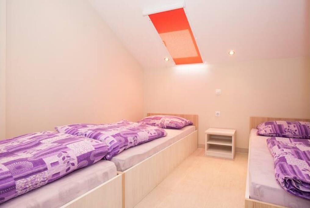 Un apartamento pequeño de una sola habitación, con dos camas, una ventana con persiana roja y una mesita de noche de madera.