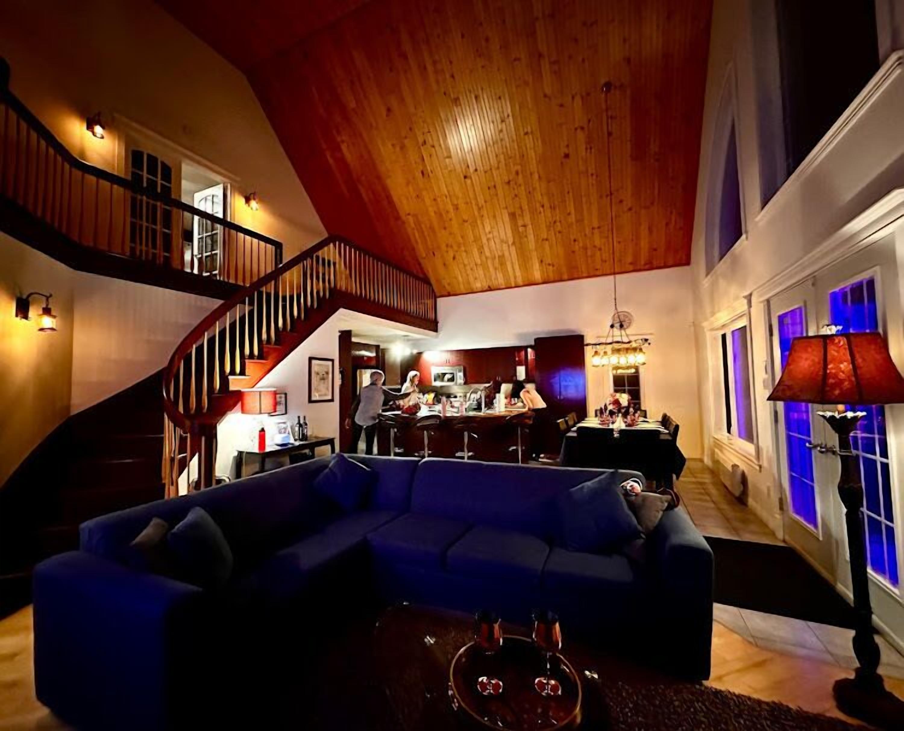 Un vaste espace de vie comprenant un grand canapé d’angle bleu, un escalier en bois et un coin repas agrémenté d’un lustre.