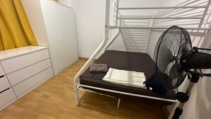 Ein Zimmer mit einem Einzelbett, einem weißen Metallgestell, einer braunen Matratze, einem weißen Nachttisch, einem weißen Kleiderschrank und einem Standventilator.
