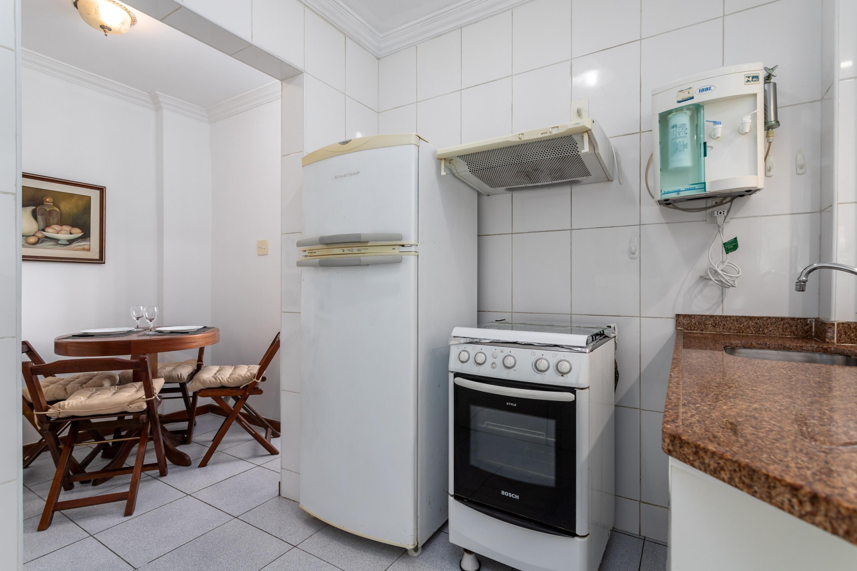 Una cocina compacta con horno Bosch, un refrigerador y dispensador de agua. Hay una mesa de comedor con sillas y un cuadro en la pared.