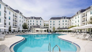 A large outdoor swimming pool surrounded by white buildings with balconies, lounge chairs, and umbrellas.