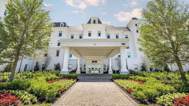 A large, white building with a central entrance and a covered walkway, surrounded by well-maintained gardens and trees.