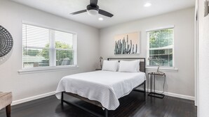 A bedroom with a bed, two windows with blinds, a ceiling fan, and a wall-mounted artwork. - Bright and Stylish Home (Dallas)