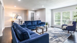 A modern living room with a sectional sofa, a coffee table, and a large window with blinds. - Bright and Stylish Home (Dallas)