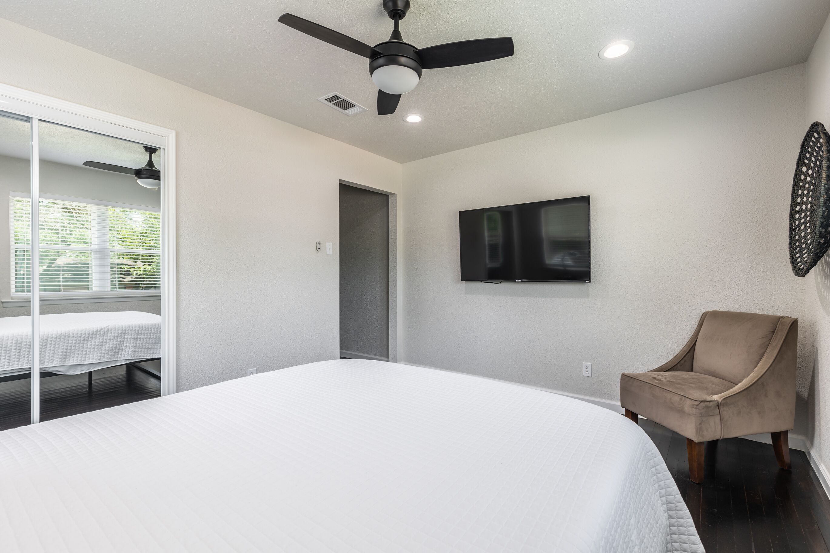 A bedroom with a large bed, a ceiling fan, a TV mounted on the wall, and a chair.