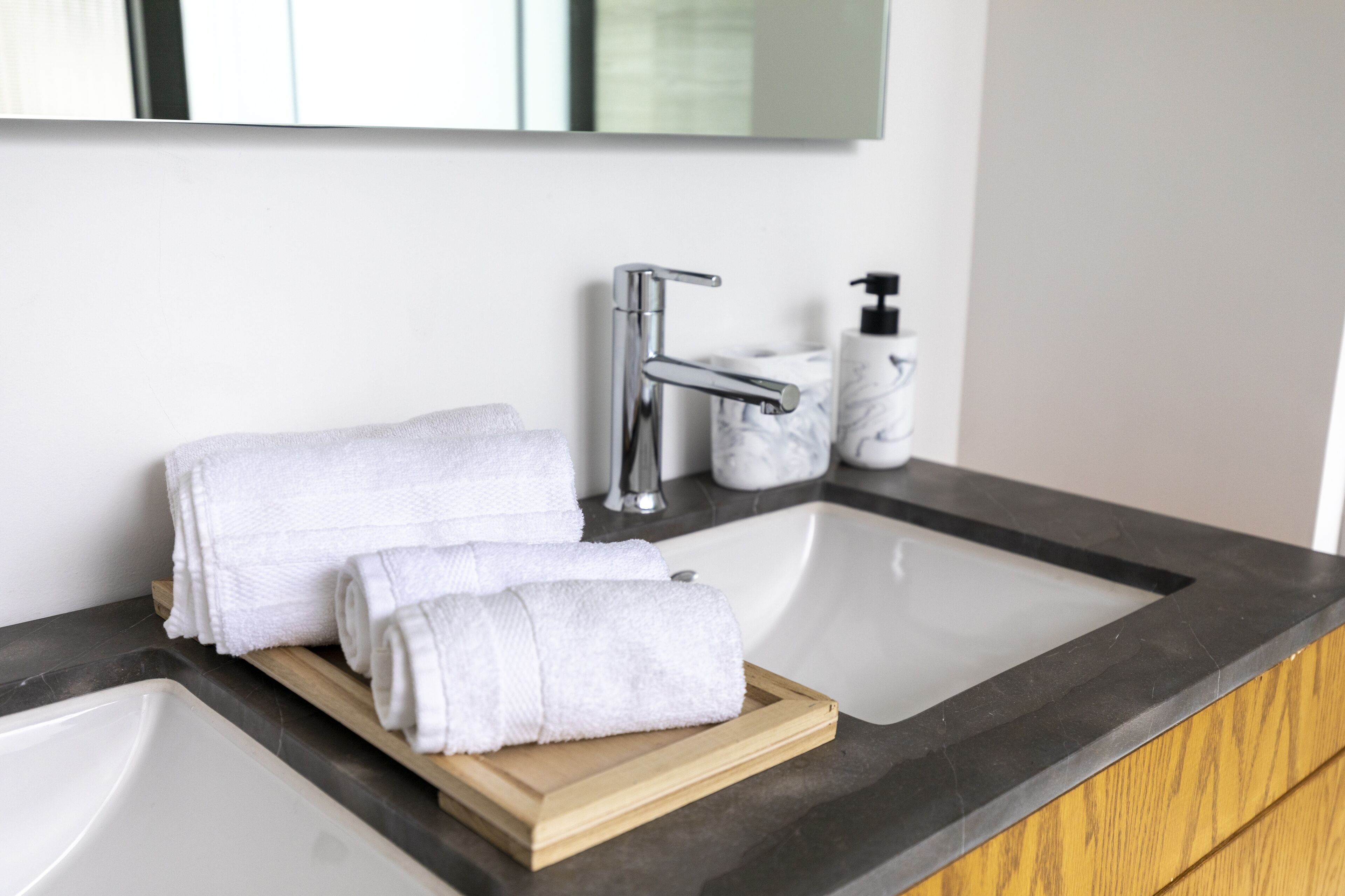 A bathroom with a white sink, a wooden tray holding rolled towels, a marble soap dispenser, and a chrome faucet.