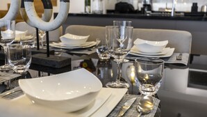 Une table à manger dressée avec de l’argenterie blanche, des verres transparents et des couverts, avec une cuisine moderne en arrière-plan.