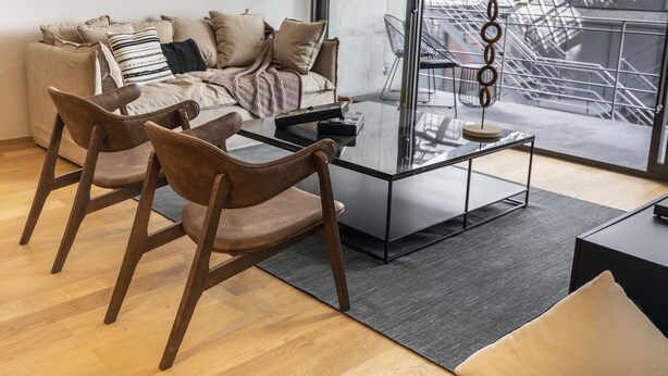 Un salon moderne avec une table basse en verre, des fauteuils en cuir, un canapé avec des coussins et une vue sur un bâtiment industriel, visible à travers de grandes fenêtres.