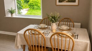 Un comedor con una mesa dispuesta para cuatro, sillas de madera, un jarrón con flores y un cuadro de paisaje enmarcado en la pared.