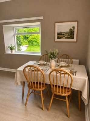A dining area with a table set for four, wooden chairs, a vase with flowers, and a framed landscape painting on the wall.