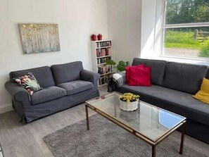 Relax in this inviting living room with two sofas, a coffee table, and a bookshelf filled with books and decorative items, offering a view of lush greenery.
