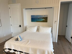 A bedroom with a bed, white walls, a painting of a coastal scene, and a door leading to another room. - In-Town Estate w/ View, Walkable to Wharfside Village, King Bed, Parking, 700sqft BRAND NEW BACKUP Electric System - No Power Outages!  Above Gallows Point (Cruz Bay)