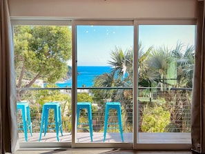 A balcony with a view of the sea, two blue stools, and a glass door. - In-Town Estate w/ View, Walkable to Wharfside Village, King Bed, Parking, 700sqft BRAND NEW BACKUP Electric System - No Power Outages! Above Gallows Point (Cruz Bay)