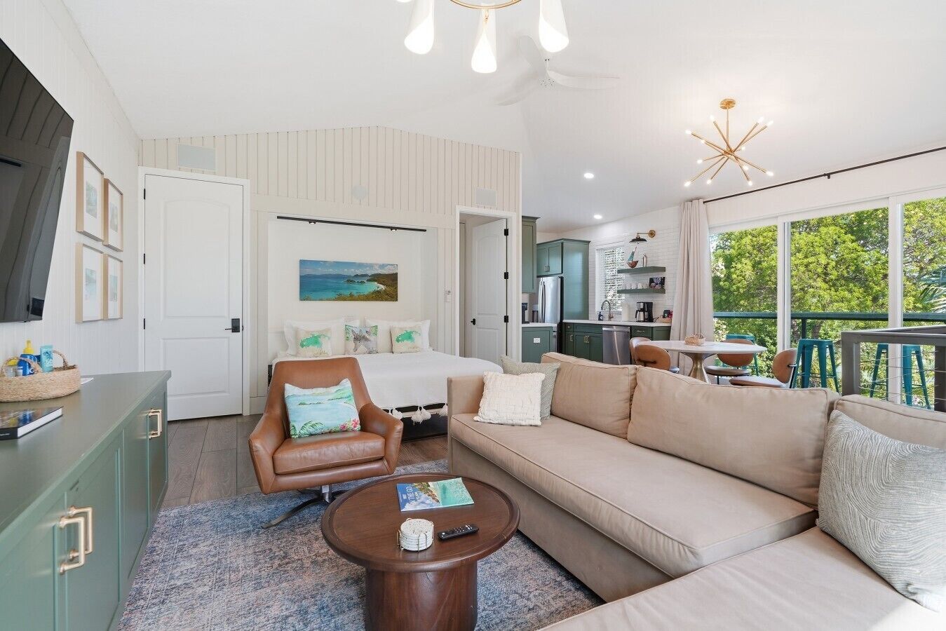 A modern living room with a sofa, armchair, coffee table, and a view of the kitchen and balcony.
