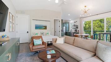 A modern living room with a sofa, armchair, coffee table, and a view of the kitchen and balcony.
