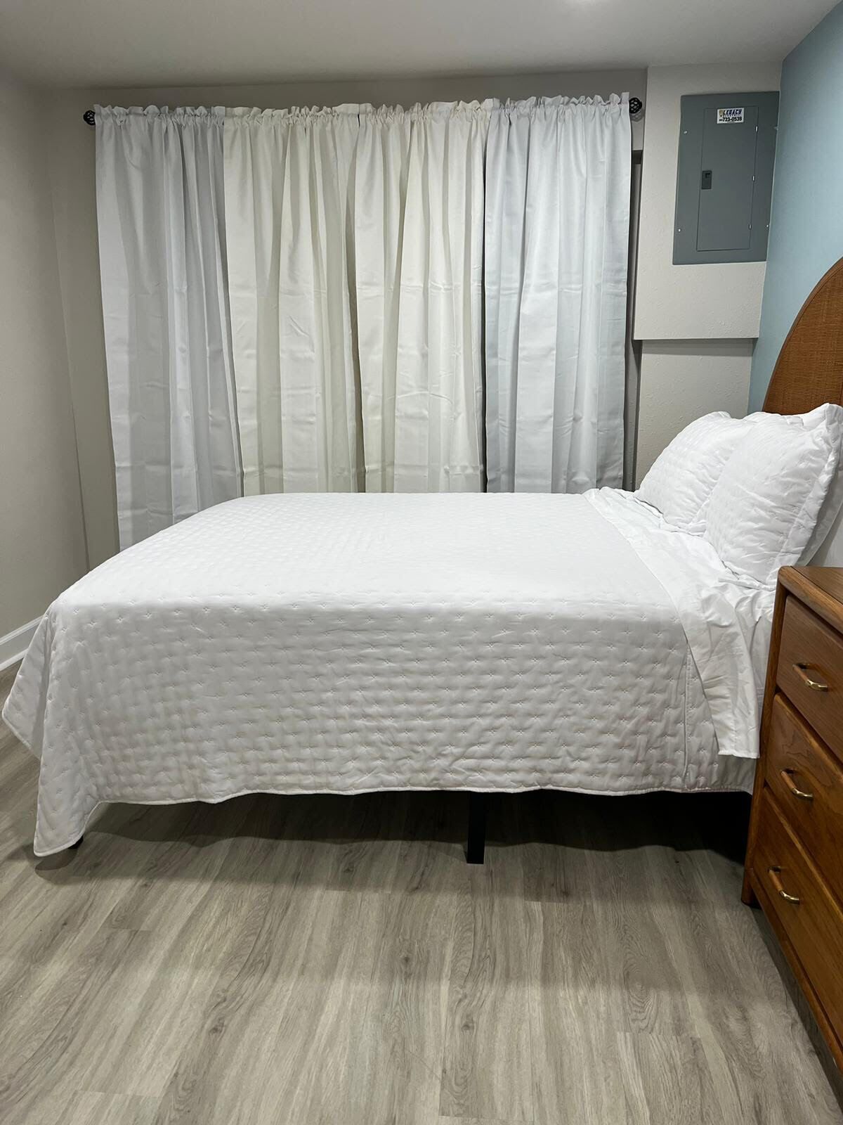 A neatly made bed with a white quilt, a wooden nightstand with drawers, and a grey wall with a utility box.
