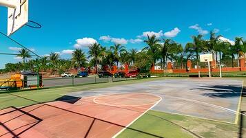 Un terrain de basket en plein air, avec un ciel dégagé, des palmiers et un bâtiment rouge en arrière-plan.