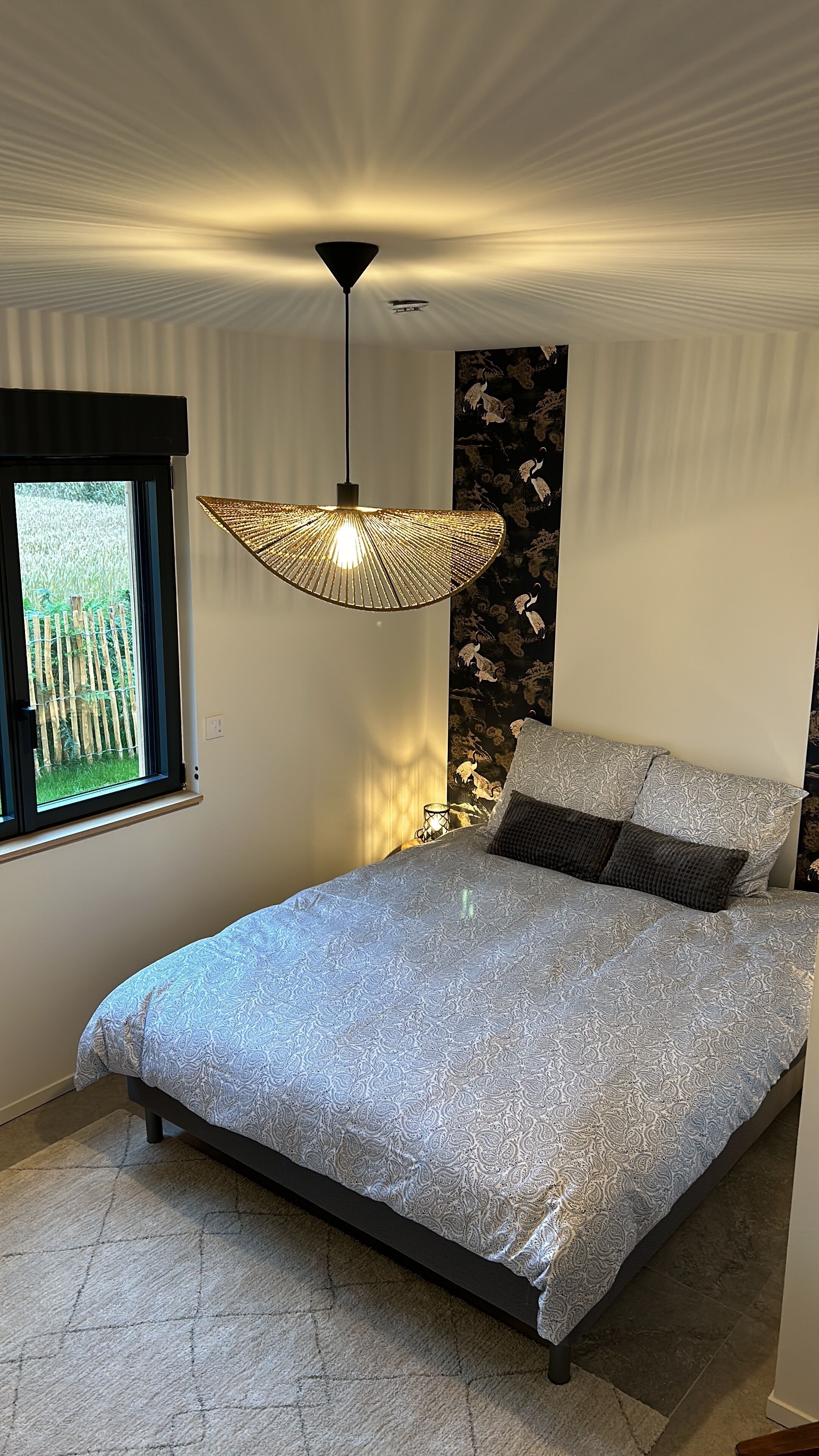A bedroom with a bed, a hanging light fixture, a window with a view of greenery, and a wall with a decorative pattern.