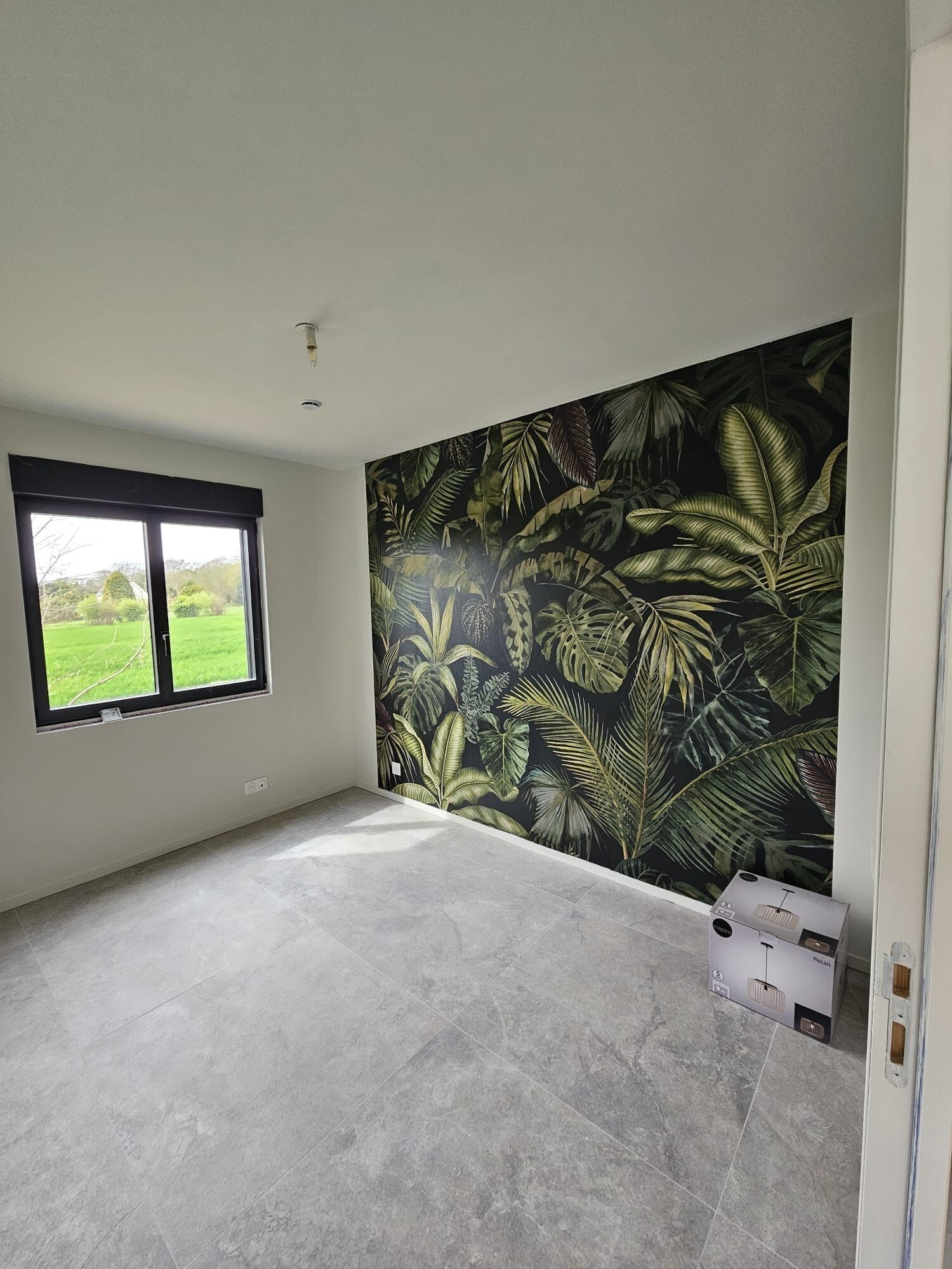 Een kamer met een groot raam, een wand met een tropisch bladmotief en een betegelde vloer.